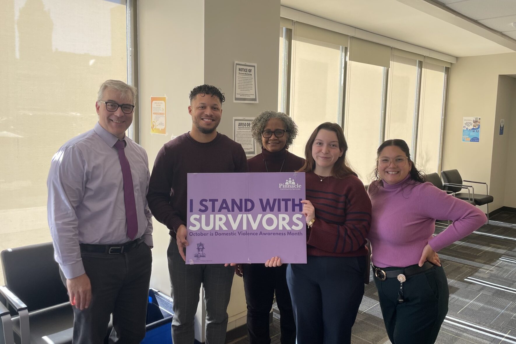 NLS team members standing in front of the window at Court St, wearing purple, holding a sign that says "I stand with survivors"