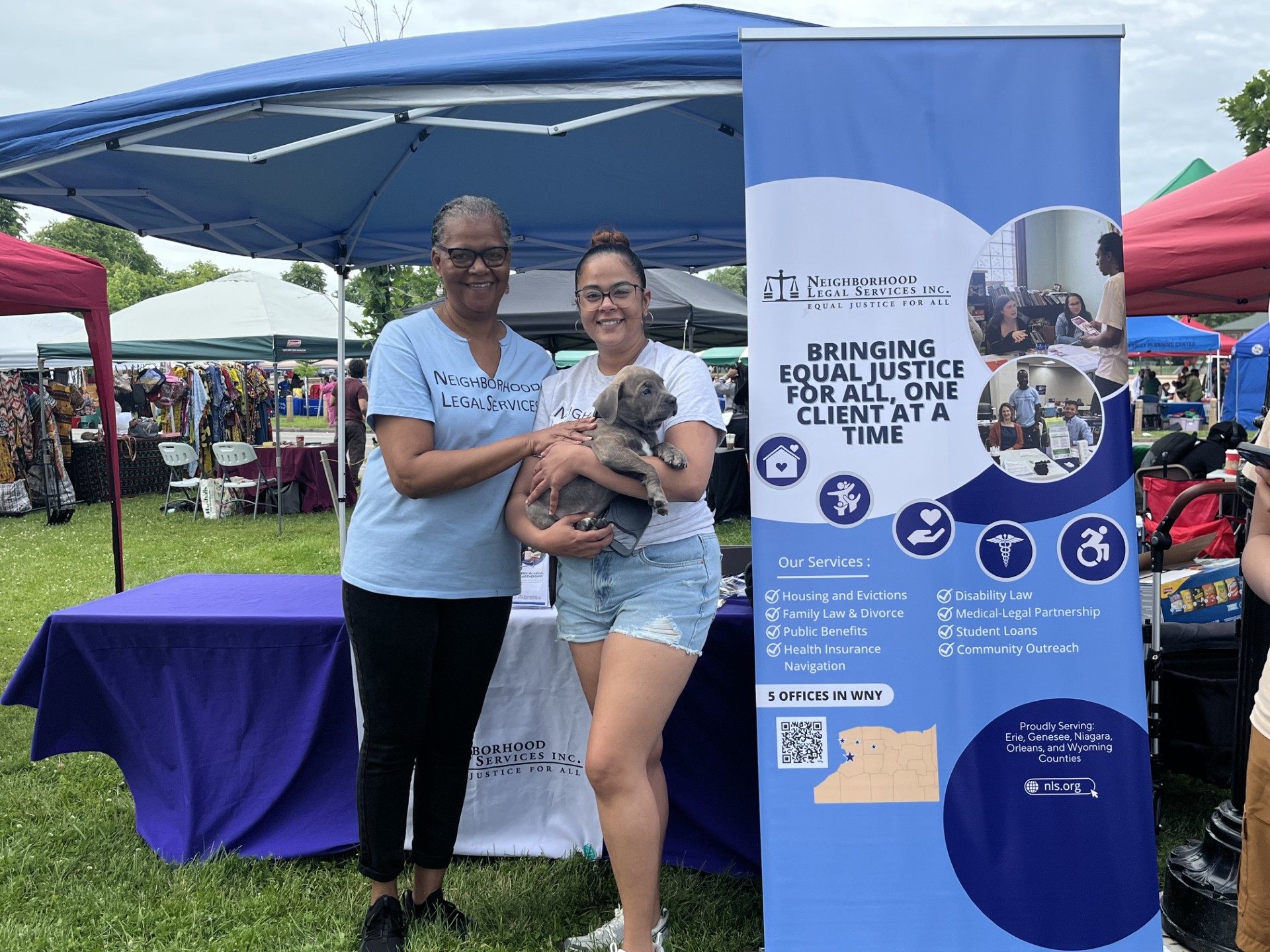 Two NLS team members standing in front of the banner sign for our organization at an outdoor tabling event, holding a small dog.